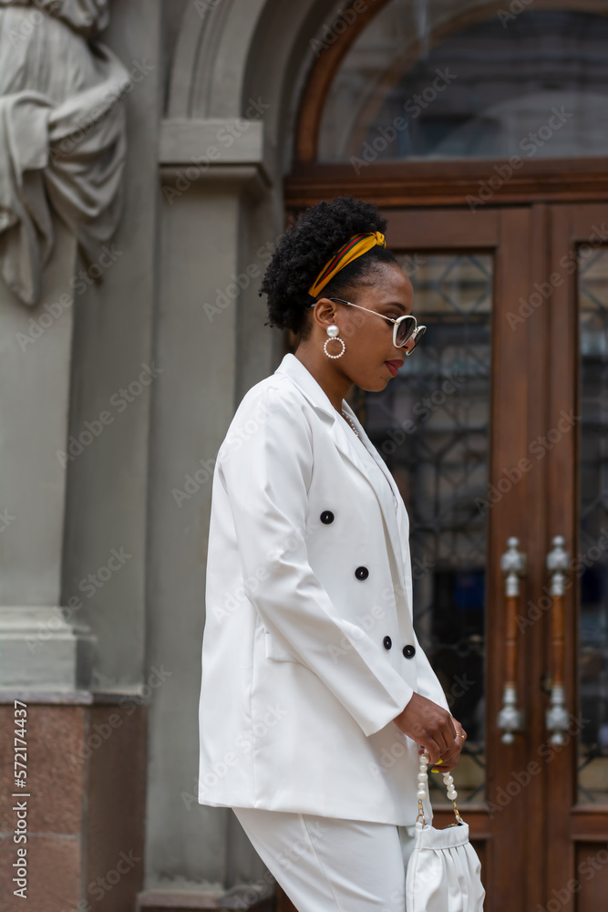 Black African-American woman in a white suit is relaxing walking in the city. Fashion portrat