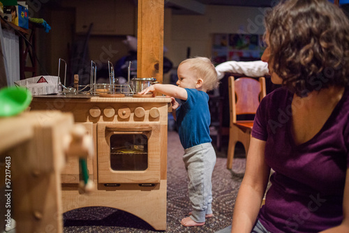 a toddler busily plays with a toy kitchen while his mother looks on