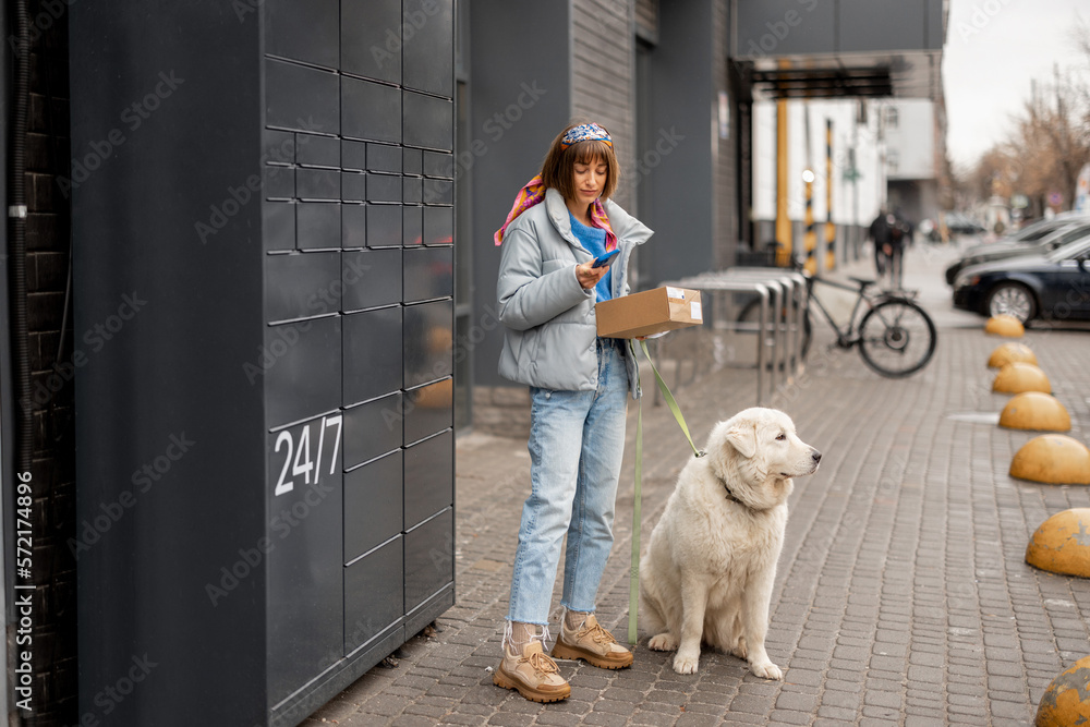 Young woman uses phone while receiving a parcel from automatic post office machine during a walk with her dog in city. Concept of modern technologies in delivery services and lifestyle Young woman uses phone while receiving a parcel from automatic post office machine during a walk with her dog in city. Concept of modern technologies in delivery services and lifestyle