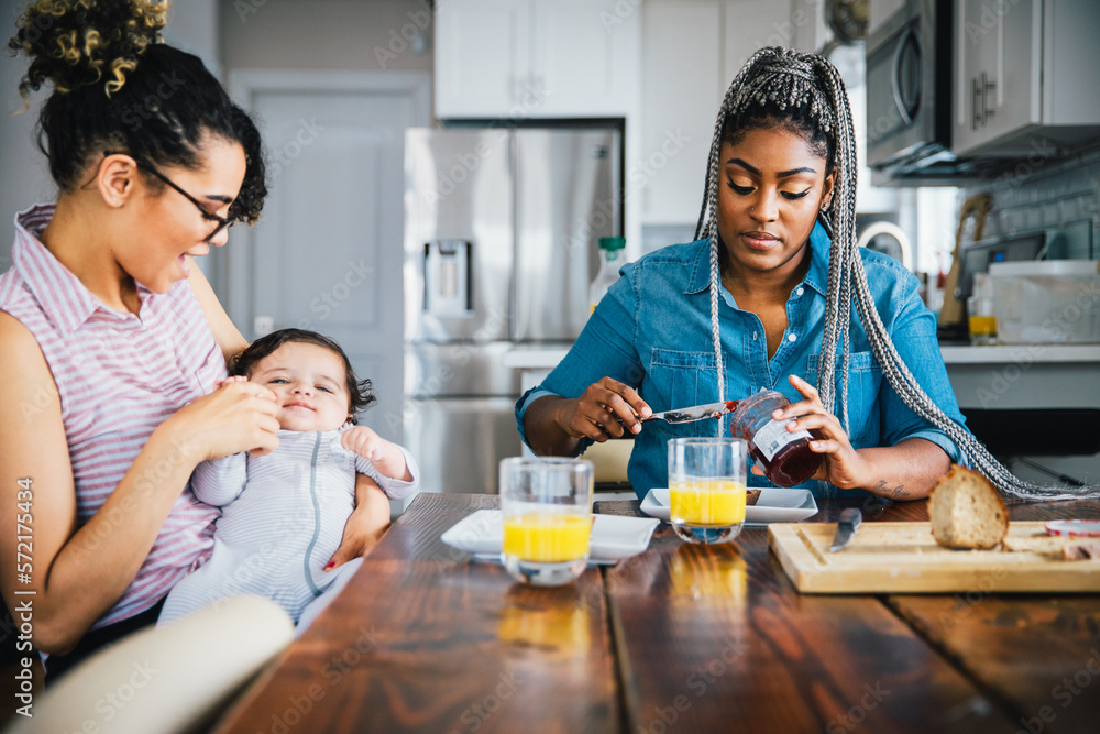 Happy mother playing with baby while girlfriend preparing breakfast at table Happy mother playing with baby while girlfriend preparing breakfast at table