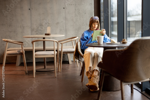 Stylish woman works on a digital tablet while sitting by the window at modern coffee shop. Concept of remote creative work online © rh2010