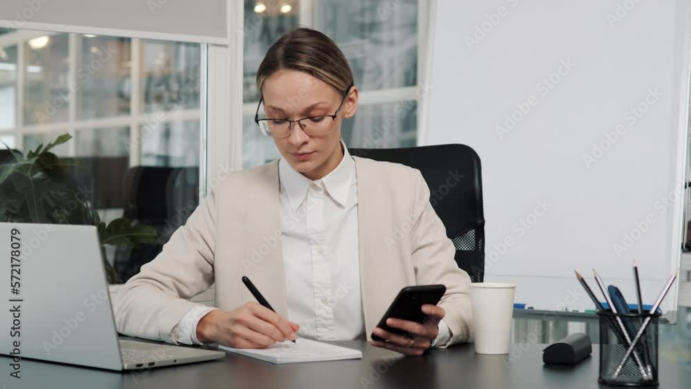 Young busy business woman office worker in eyeglasses working on project paperwork using mobile phone. Smart female manager making budget writing tasks from cellphone working alone in office. Young busy business woman office worker in eyeglasses working on project paperwork using mobile phone. Smart female manager making budget writing tasks from cellphone working alone in office.