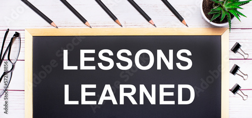 On a light wooden table - a board with the text LESSONS LEARNED, pencils, plants, glasses and paper clips. Business concept