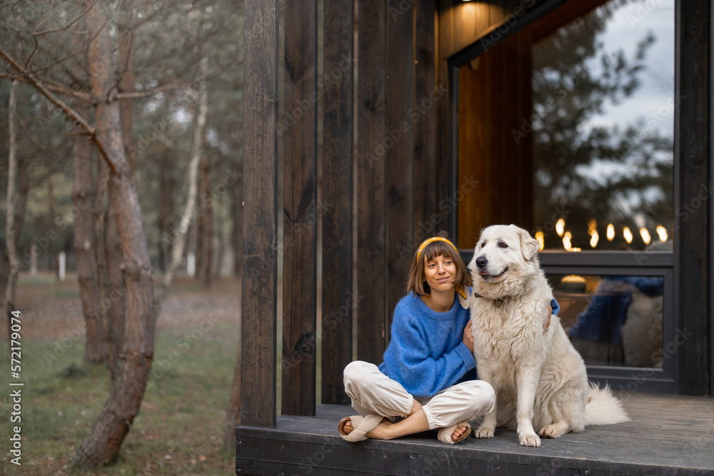 © rh2010 - Young woman sits with her dog on porch of a wooden house in pine forest, enjoying nature while resting in cottage at countryside
