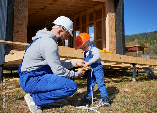 Father with toddler son building wooden frame house. Male builder and kid playing with tape measure on construction site, wearing helmet and blue overalls on sunny day. Carpentry and family concept.