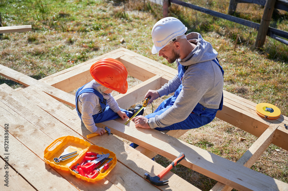 Father with toddler son building wooden frame house. Boy helping his daddy, playing with tape measure on construction site, wearing helmet and blue overalls on sunny day. Carpentry and family concept.