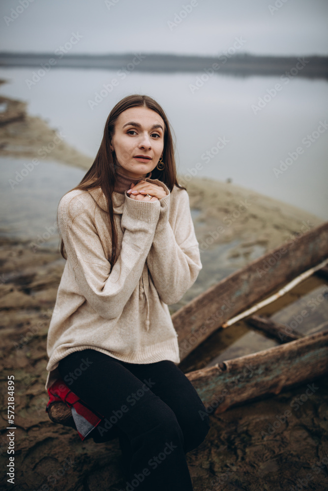 beautiful woman on the shore of a foggy lake, cold weather. A wooden boat on which a woman sits. a girl in a warm beige sweater, romantic photos against the background of a foggy lake