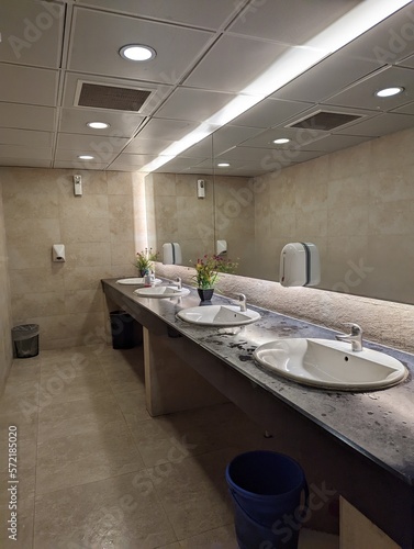 A large washroom with a lot of basins on a row with a big mirror hanging on the wall, a blue colored dustbin placed below, also with great lightning all around the surroundings. 