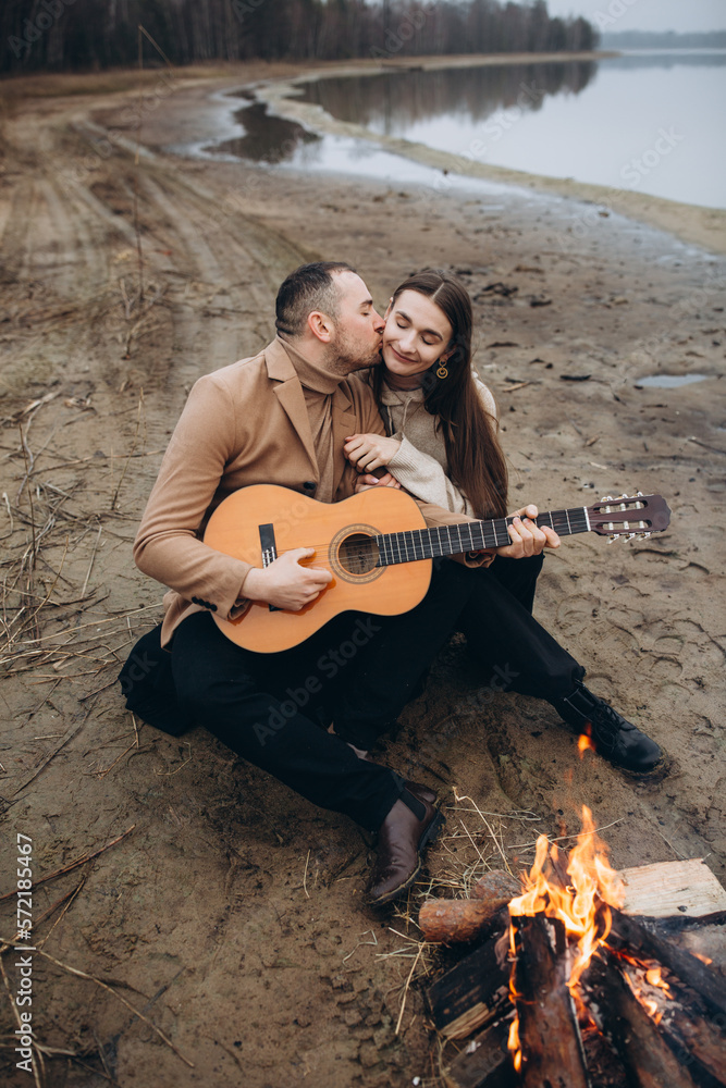 Fototapeta premium a man on the shore of a lake near a fire plays the guitar for a girl. foggy cold weather. beige men's coat. bonfire on the shore of the reservoir. photo in beige tones. romantic photo with a guitar