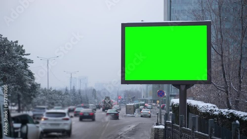 4k Mockup poster stands on street with passing cars in city spbas. Lot of transport raffic is driving on road and green screen lightbox standing outdoors in snowy winter weather. Vertical billboard is photo