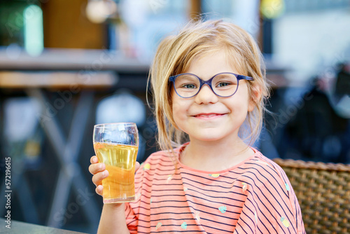Adorable little girl having breakfast and drinking juice at resort restaurant. Happy preschool child with glasses holding glass with apple juice