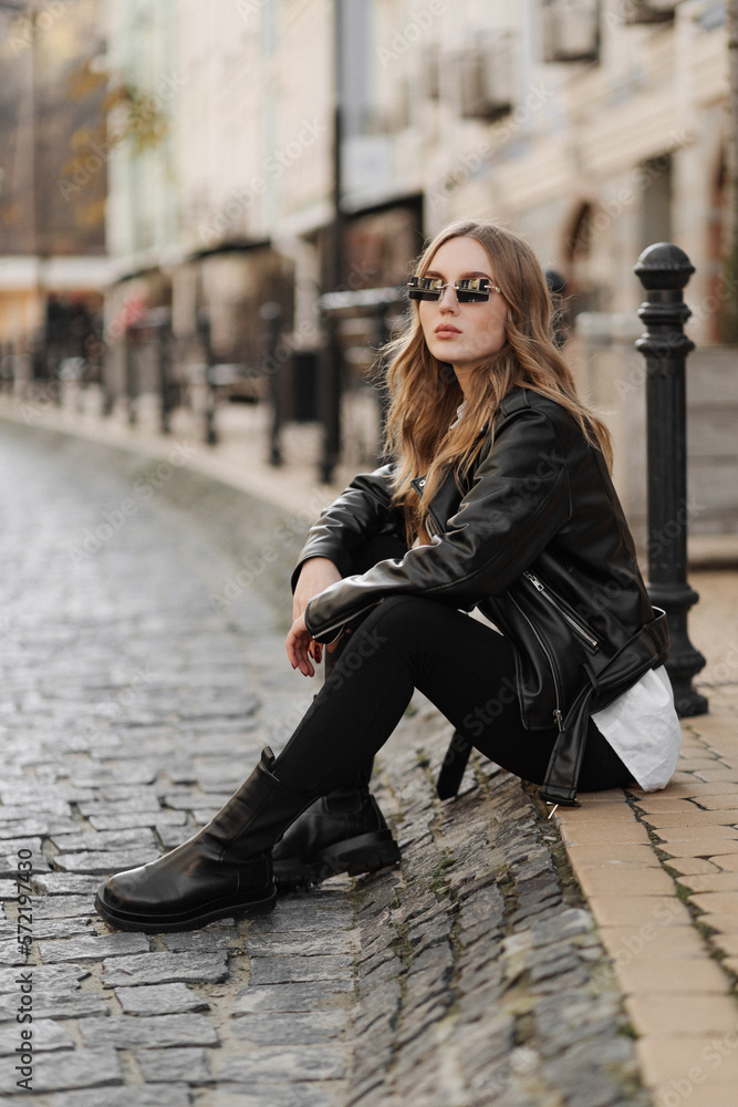 Fashionable blonde woman model with black leather jacket and style sunglasses sitting on a floor at the city Fashionable blonde woman model with black leather jacket and style sunglasses sitting on a floor at the city