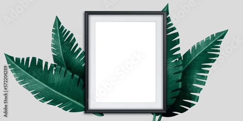 An empty white photo frame and green leaves are on a gray concrete background. Floral layout. Flat lay. Top view. Copy space.