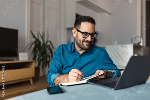 Adult man working over the laptop, taking notes in the notebook, sitting on the home floor.