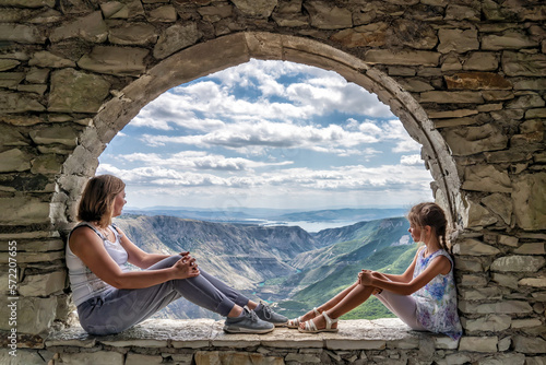 Happy woman with her daughter sits on stone window with breathtaking view of valley of Sulak Canyon and reservoir surrounded by mountain ranges ahead