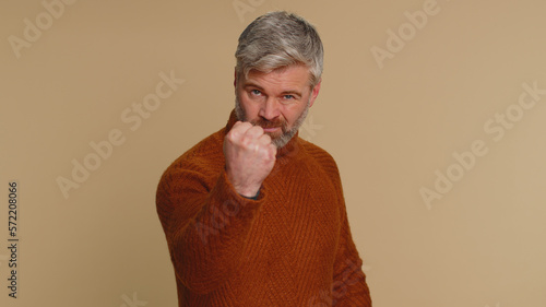 Aggressive angry middle-aged man trying to fight at camera, shaking fist, boxing with expression, punishment, disappointment, rage, quarrel. Senior mature guy isolated alone on beige studio background