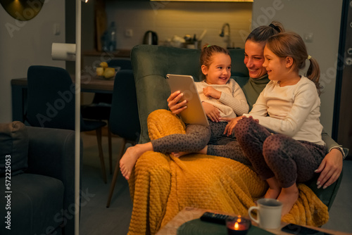 Mother and daughters are comfortable on an armchair looking at videos on a tablet, they are happy and are laughing