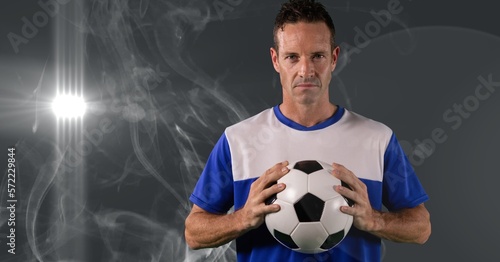 Caucasian male soccer player holding football against smoke effect and light spot on grey background