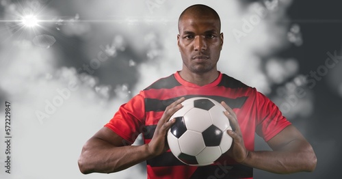 African american male soccer player holding football against light spot on grey background