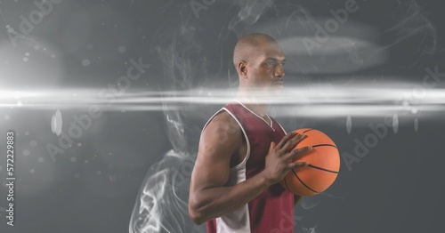 African american male basketball player holding ball against smoke effect on grey background © vectorfusionart