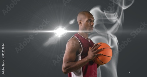 African american male basketball player holding ball against smoke and light spot on grey background