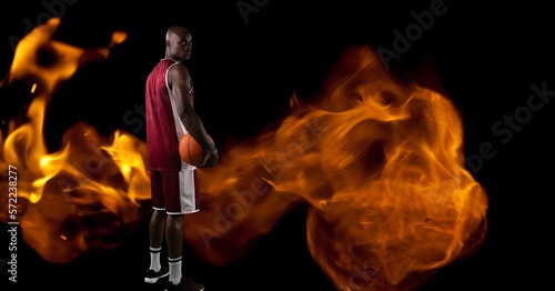 Composition of male basketball player standing holding ball over flames on black background