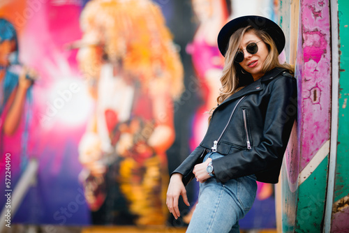 The young stylish woman in black sunglasses, hat, leather jacket wearing braces posing at the amusement park's multicolored wall   © Natalia