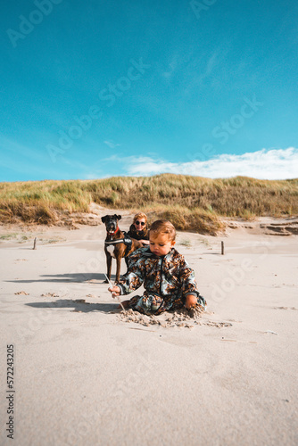 Kleinkind sitz am Strand im Vordergrund, dahinter hocken Mutter und Hund und schauen zu der Tochter die im Sand spielt