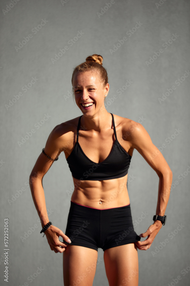 Fototapeta premium Healthy young sportswoman doing fitness. Sportswoman raises her leg, does functional training outdoors on a bright sunny day, smiles contentedly, dressed in a sports uniform