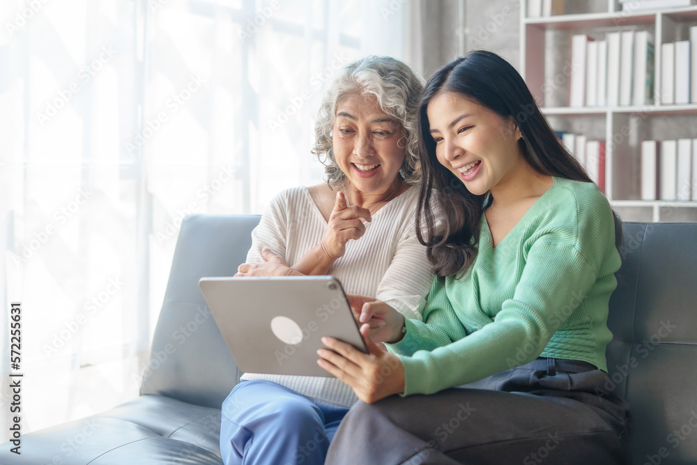 60s asian mother elderly sitting on sofa with young asia female daughter together in living room. watch movies series online or shopping, using tablet computer