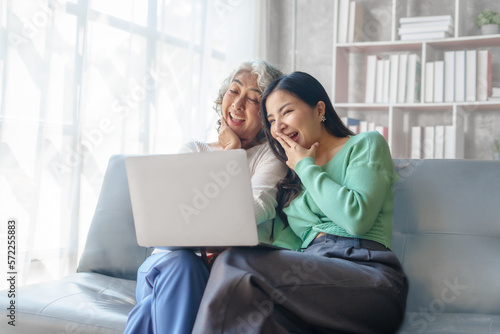 60s asian mother elderly sitting on sofa with young asia female daughter together in living room. watch movies series online or shopping, using laptop computer