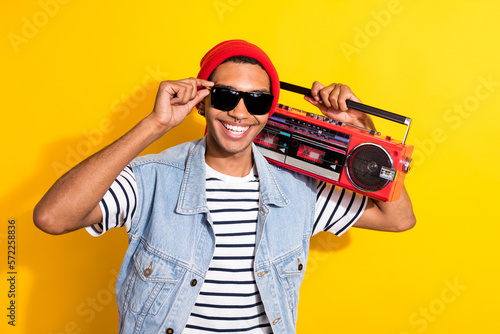 Photo of cheerful carefree man arm touch sunglass carry boombox toothy smile isolated on yellow color background