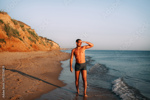 A slender, tanned guy in blue swimming trunks walks along the sandy shore of the ocean and looks into the distance, on a bright sunny day. Hills are visible in the distance.