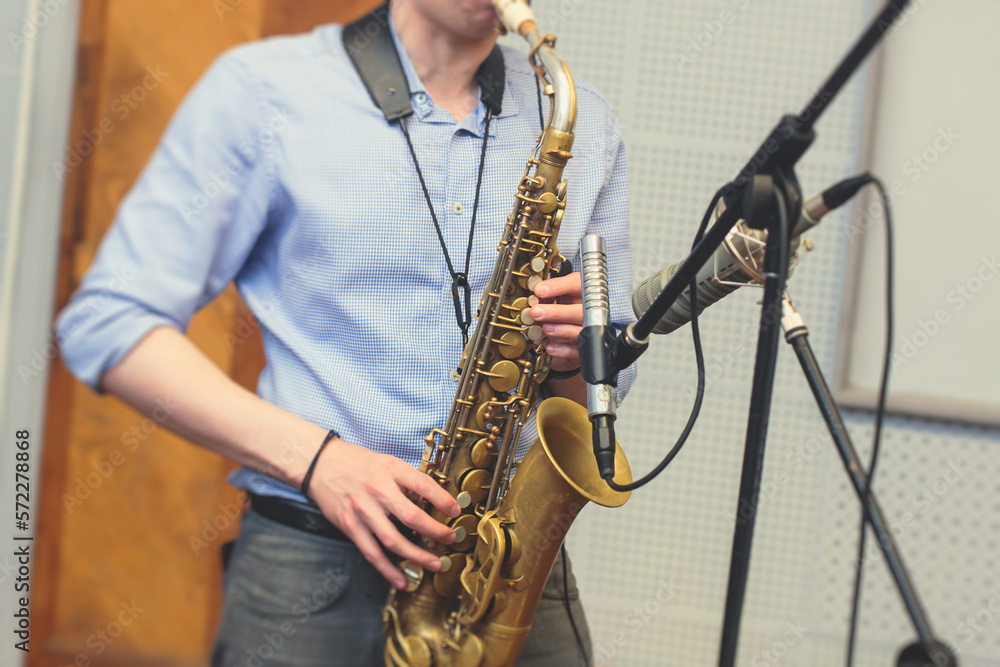 Naklejka premium View of a saxophone player in headphones during rehearsal, recording sound for new album song at studio, saxophonist musician in front of microphone with musical band orchestra, music production