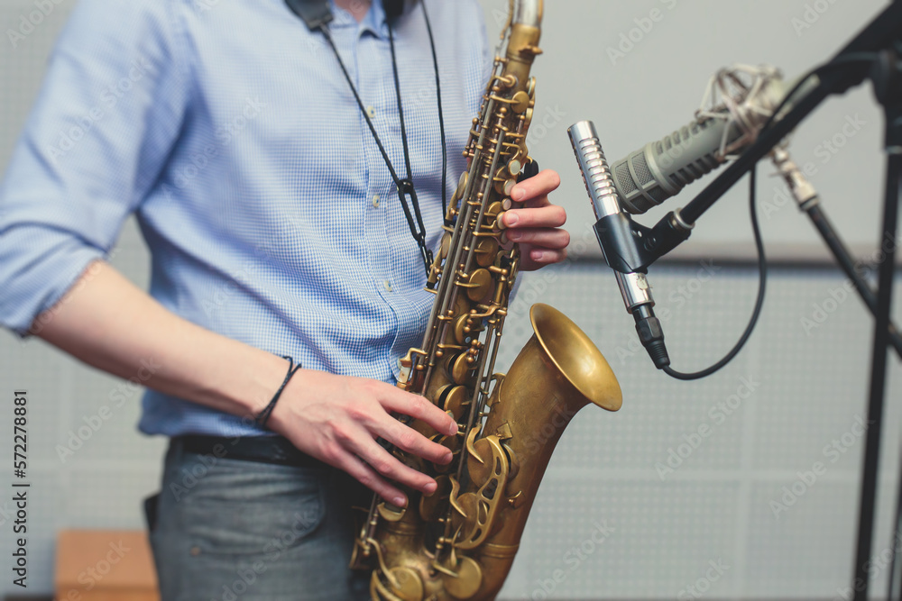 Naklejka premium View of a saxophone player in headphones during rehearsal, recording sound for new album song at studio, saxophonist musician in front of microphone with musical band orchestra, music production