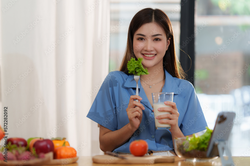 Portrait of a young cheerful asian woman drinking milk in kitchen room with healthy raw food. Vegetarianism, wellbeing and healthy lifestyle concept.