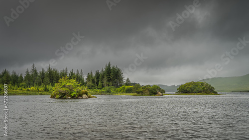 Small rocky islands covered by bushes and trees on Ballynahinch Lake. Pine forest with dark storm sky in background. Connemara, County Galway photo
