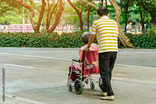 Back view of Asian elderly man walking with disabled elderly woman sitting in wheelchair outdoors wearing medical masks. Man pushes old lady in wheelchair through park. Healthy strong medical concept.