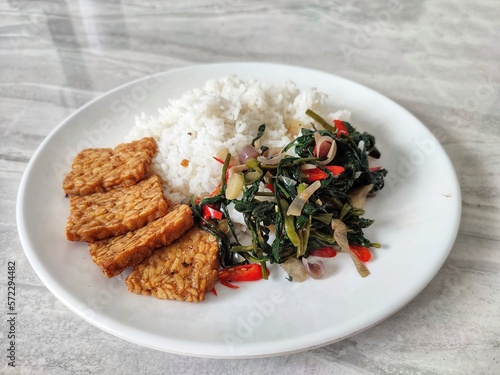 A simple menu consisting of rice, stir-fried kale or tumis kangkung and fried tempeh or tempe goreng on a white plate on a black and white carpet. Indonesian simple menu photo