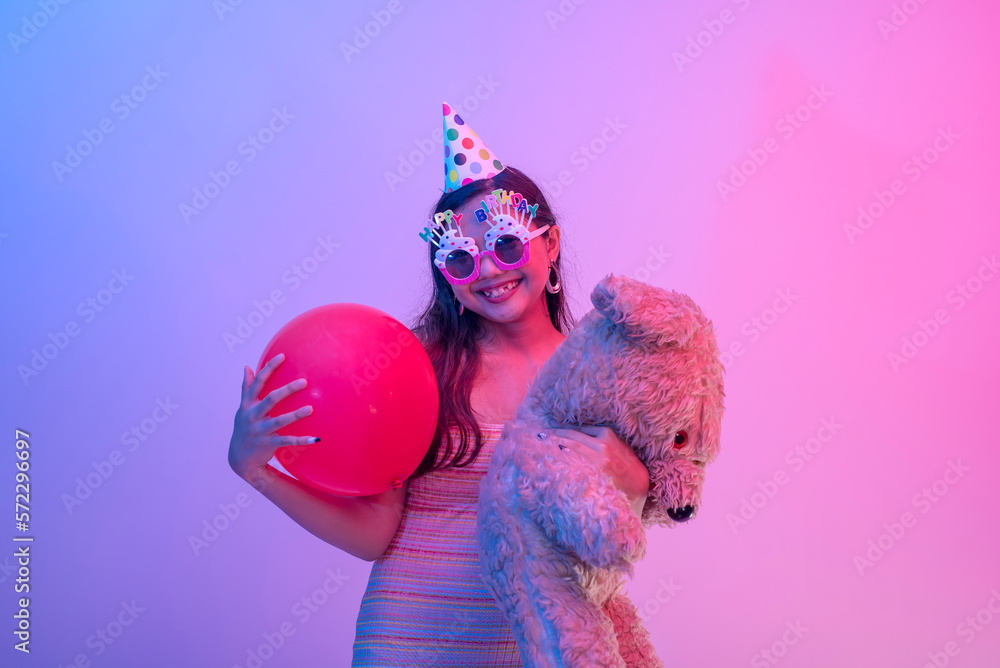 A young female birthday celebrant wearing a party hat and novelty glasses and holding a red balloon and a teddy bear. An asian lady partying. Lit with blue and pink neon colors.
