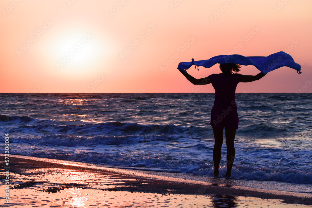 Silhouette of a girl with a blue shawl on the seashore at sunrise, back view