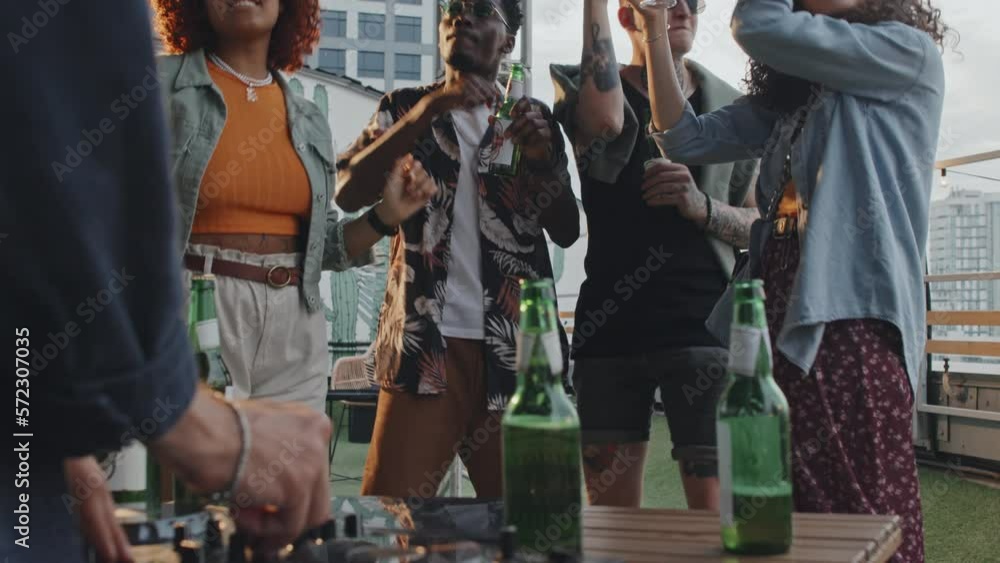 Selective focus of young ethnically diverse people enjoying summer evening party on rooftop with DJ mixing music on console Selective focus of young ethnically diverse people enjoying summer evening party on rooftop with DJ mixing music on console