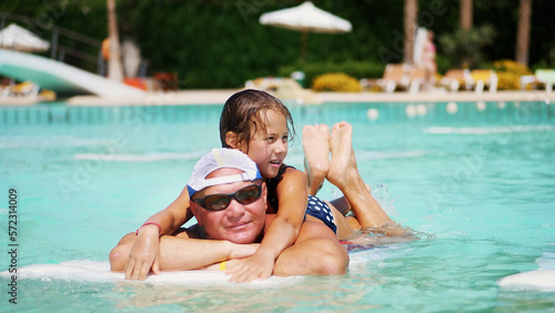 man in sun glasses, father and daughter, kid girl, playing in the pool water, having fun together. Happy family relaxing by the swimming pool on a hot summer day at water park. Summer vacation concept