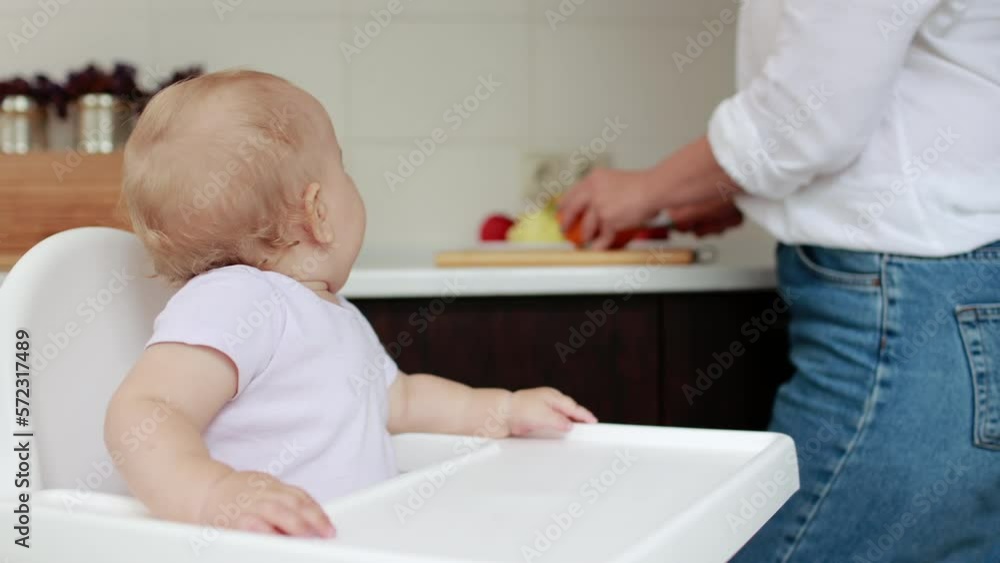 First baby food. Mom cutting fresh raw organic fruits for her cute baby girl boy, who sitting in highchair, at home kitchen.Bio fruits as first solid food for infant. Healthy nutrition snack for kids. First baby food. Mom cutting fresh raw organic fruits for her cute baby girl boy, who sitting in highchair, at home kitchen.Bio fruits as first solid food for infant. Healthy nutrition snack for kids.