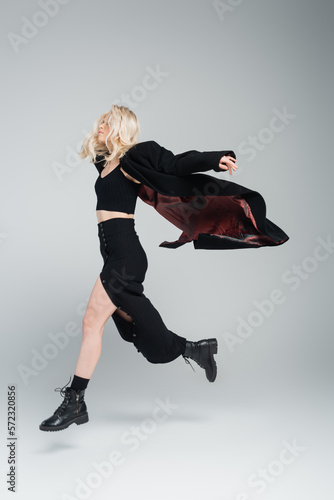 full length of blonde and young woman in stylish black outfit jumping on grey.