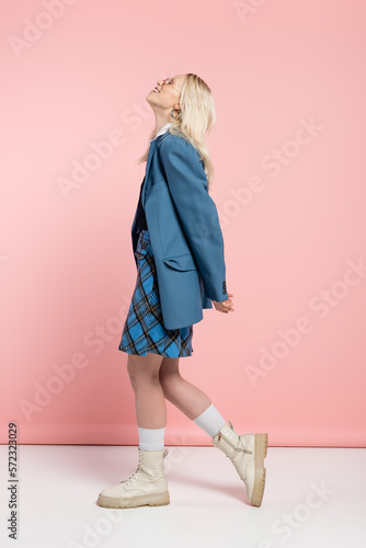 Fotografie full length of smiling blonde woman in blue outfit and trendy sunglasses posing with raised head on pink