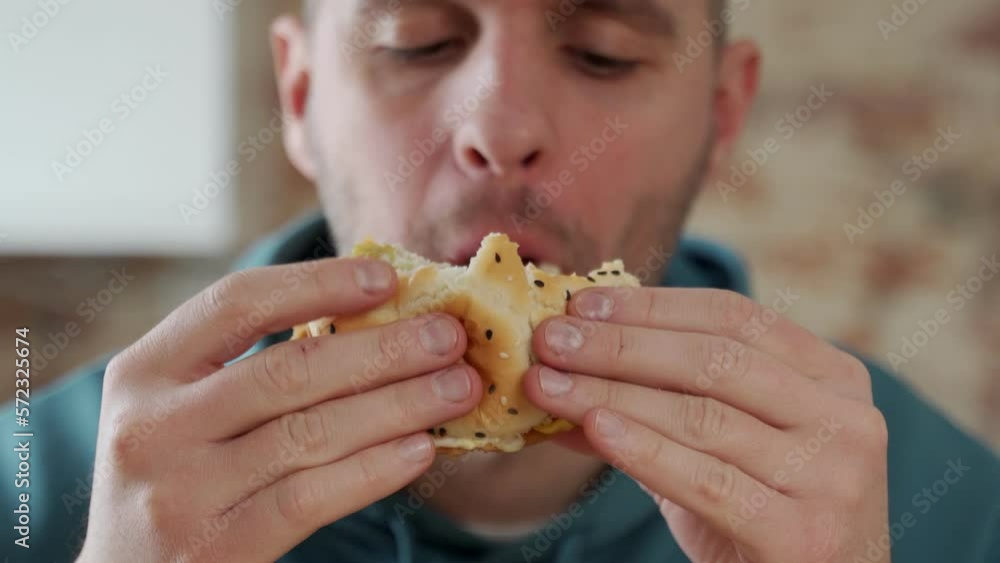 Close-up of a young man greedily eating a delicious hamburger, feeling very hungry, consuming fast food Close-up of a young man greedily eating a delicious hamburger, feeling very hungry, consuming fast food
