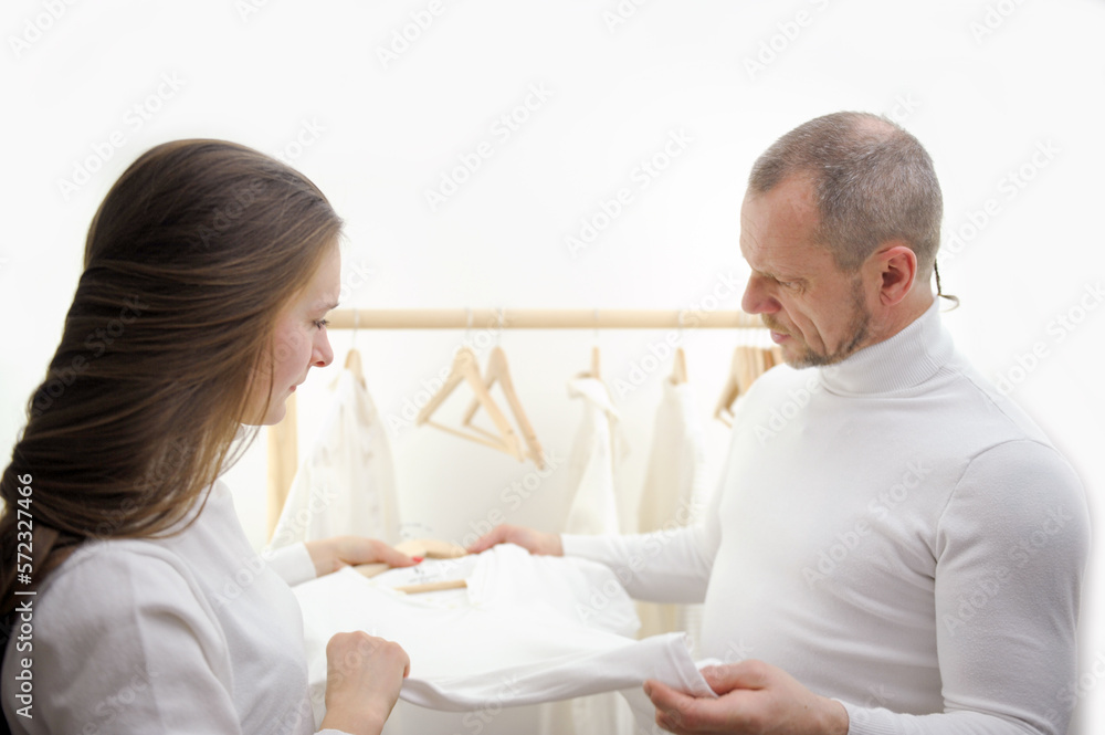 Fototapeta premium clothing store young girl seller offers a white t-shirt to a man man frowning seriously examines her beard wrinkles on her face tenderness of a young girl to a man 50-60 years old girls 20-25 studio