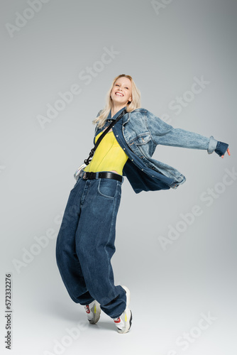 full length of happy young woman in blue denim outfit and stylish sneakers posing on grey.
