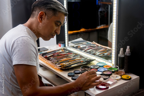 young man with dark skin applying makeup to himself in front of a mirror with a table full of makeup and brushes to change and improve his facial aesthetics photo